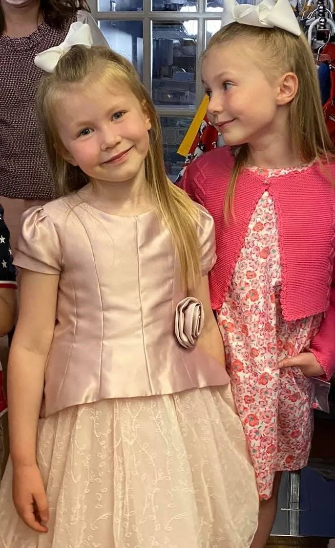 Two girls in party dresses, one blush satin and one pink floral with a cardigan