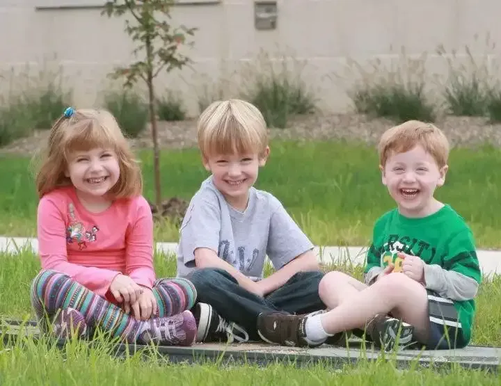 Three smiling young children sitting on the grass in casual play clothes