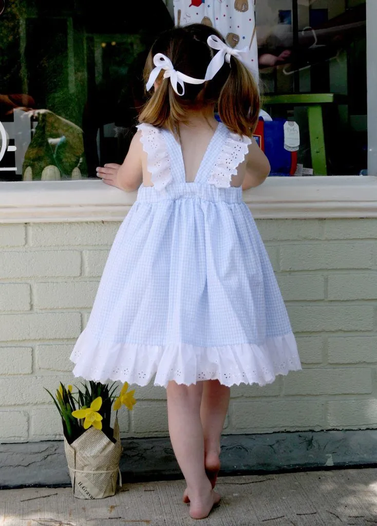 Toddler girl in a blue gingham pinafore dress with eyelet trim looking into a shop window